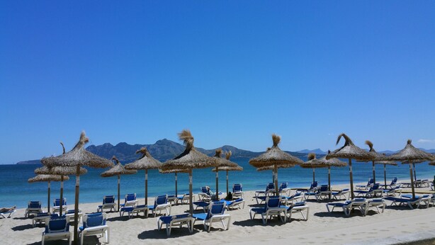 En strand med rader av solstolar och parasoller med halmtak, en klarblå himmel och berg i bakgrunden.