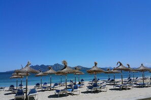 A beach with rows of sun loungers and thatched umbrellas, a clear blue sky, and mountains in the background. - Beachfront apartment in Puerto de Pollensa (Port de Pollença)