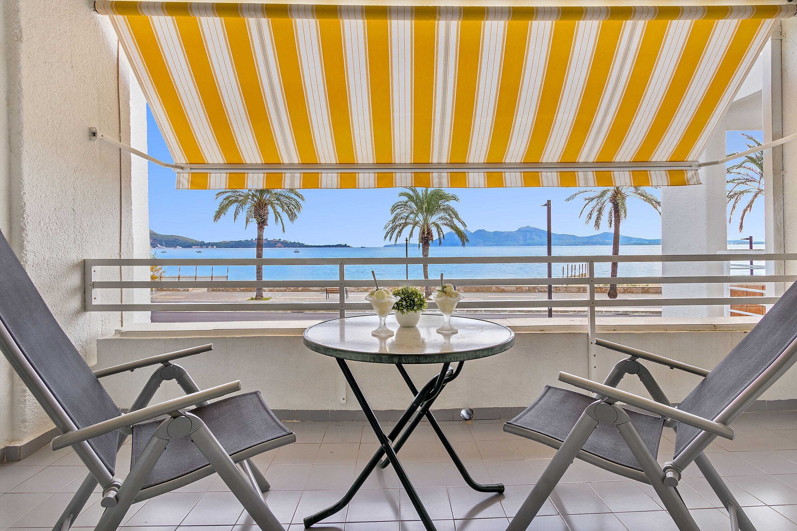 A balcony with a yellow-striped awning, two lounge chairs, a small table with a vase and two glasses, and a view of the sea and palm trees.
