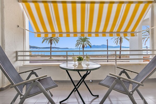 Un balcón con toldo a rayas amarillas, dos sillones de descanso, una mesita con un jarrón y dos vasos, y vistas al mar y palmeras.