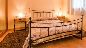 Een slaapkamer met een metalen bedframe, een bloemenprint beddeken, twee nachtkastjes met lampen, en een groot raam met gordijnen.
