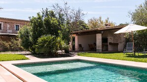 Una piscina rectangular con borde de azulejos, rodeada de césped y un edificio de piedra con un porche cubierto.