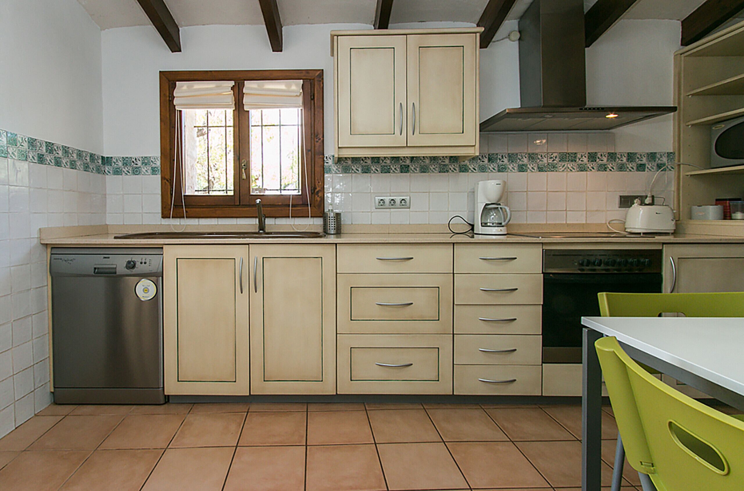 Une cuisine avec des meubles de couleur beige, un lave-vaisselle, une fenêtre avec des stores et une table avec une chaise.