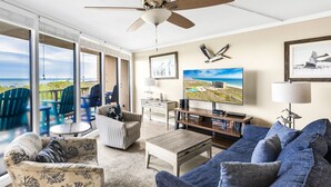 A spacious living room with a large sofa, a coffee table, and a television. There is a balcony with outdoor seating and a view of the ocean. - Sea Gull #103: Beachfront 2 Bedroom 2 Bathroom With Spectacular Views and 24 Hour Management (Port Aransas)
