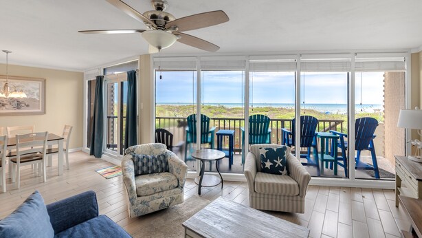 A spacious living room with a ceiling fan, a dining area, and a balcony with blue chairs and a table. - Sea Gull #103: Beachfront 2 Bedroom 2 Bathroom With Spectacular Views and 24 Hour Management (Port Aransas)