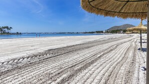 A beach with a thatched umbrella, sandy beach, and a distant mountain. - 1st line apartment Alcudia beach CAB (Puerto de Alcudia)