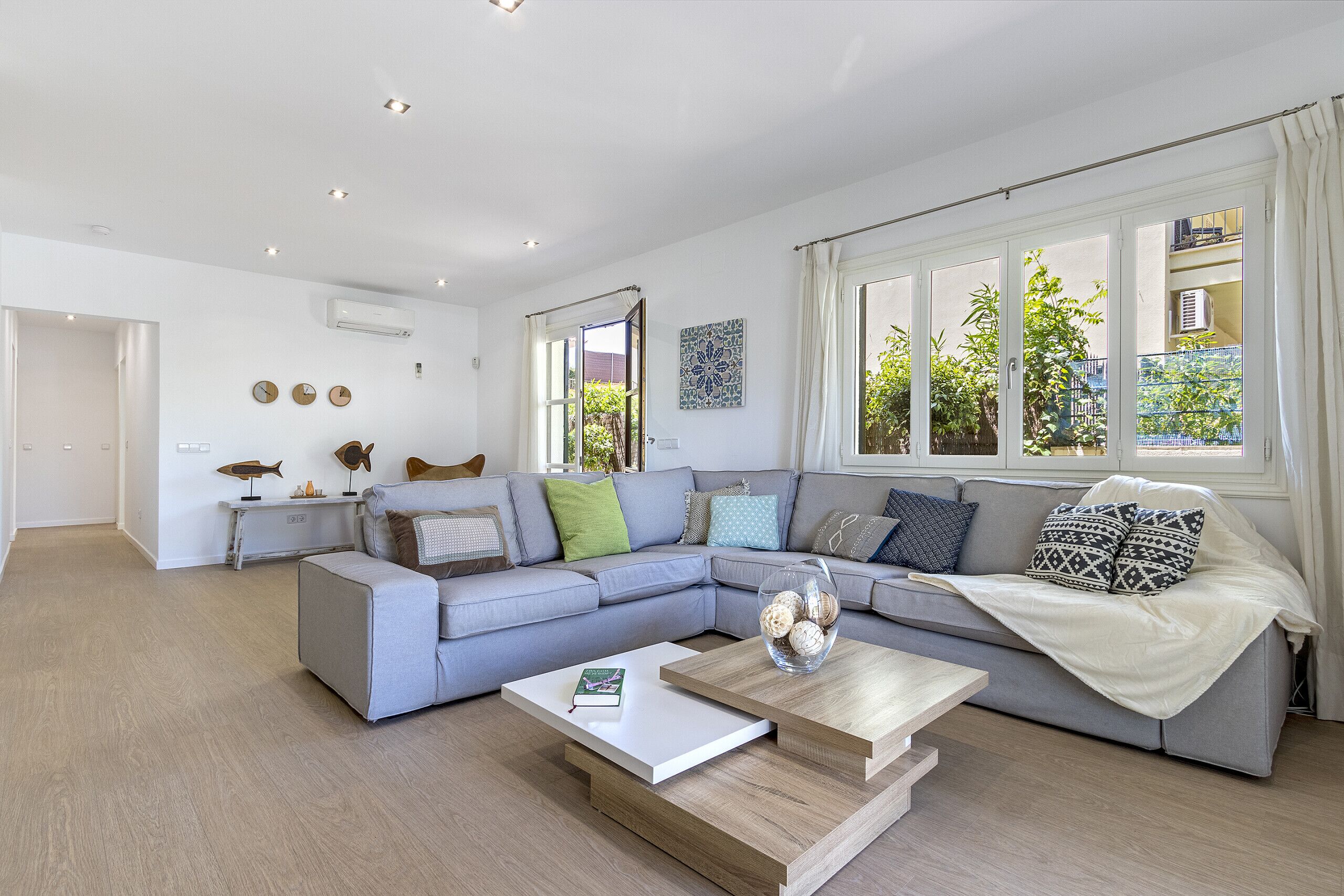 Un salon moderne avec un canapé d’angle gris, une table basse en bois et une vue sur la verdure à l’extérieur, grâce à de grandes fenêtres.