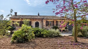A stone house with a tiled roof, surrounded by a garden with various plants and a tree with pink flowers. - Country house with amazing swimming pool and garden in a beautiful rural setting (Campanet)