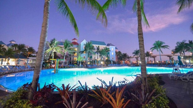 A resort pool area with palm trees, lounge chairs, and a building with a red roof.