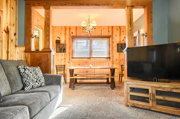 A cozy living room with a wooden interior, a sofa with a patterned pillow, a wooden dining table, and a flat-screen TV on a wooden cabinet. - Black Barrel Lodge (Lead)