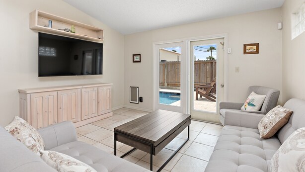 A living room with a sofa, a coffee table, a TV mounted on the wall, and a sliding glass door leading to an outdoor area. - 2-min Walk to Beach, Private House with Heated Pool (South Padre Island)