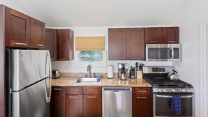 A modern kitchen with dark wooden cabinets, stainless steel appliances, a dishwasher, a sink, and a window with a blind. - Rex- East Hampton Cottage- 5 stars (East Hampton)