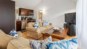 A living room with a beige sofa, a coffee table, and a TV. There is a kitchen in the background with wooden cabinets and a microwave. - Rex- East Hampton Cottage- 5 stars (East Hampton)