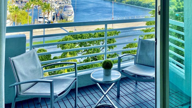 Un balcon avec deux chaises et une petite table, donnant sur une marina où sont amarrés des bateaux, ainsi que sur une rivière.