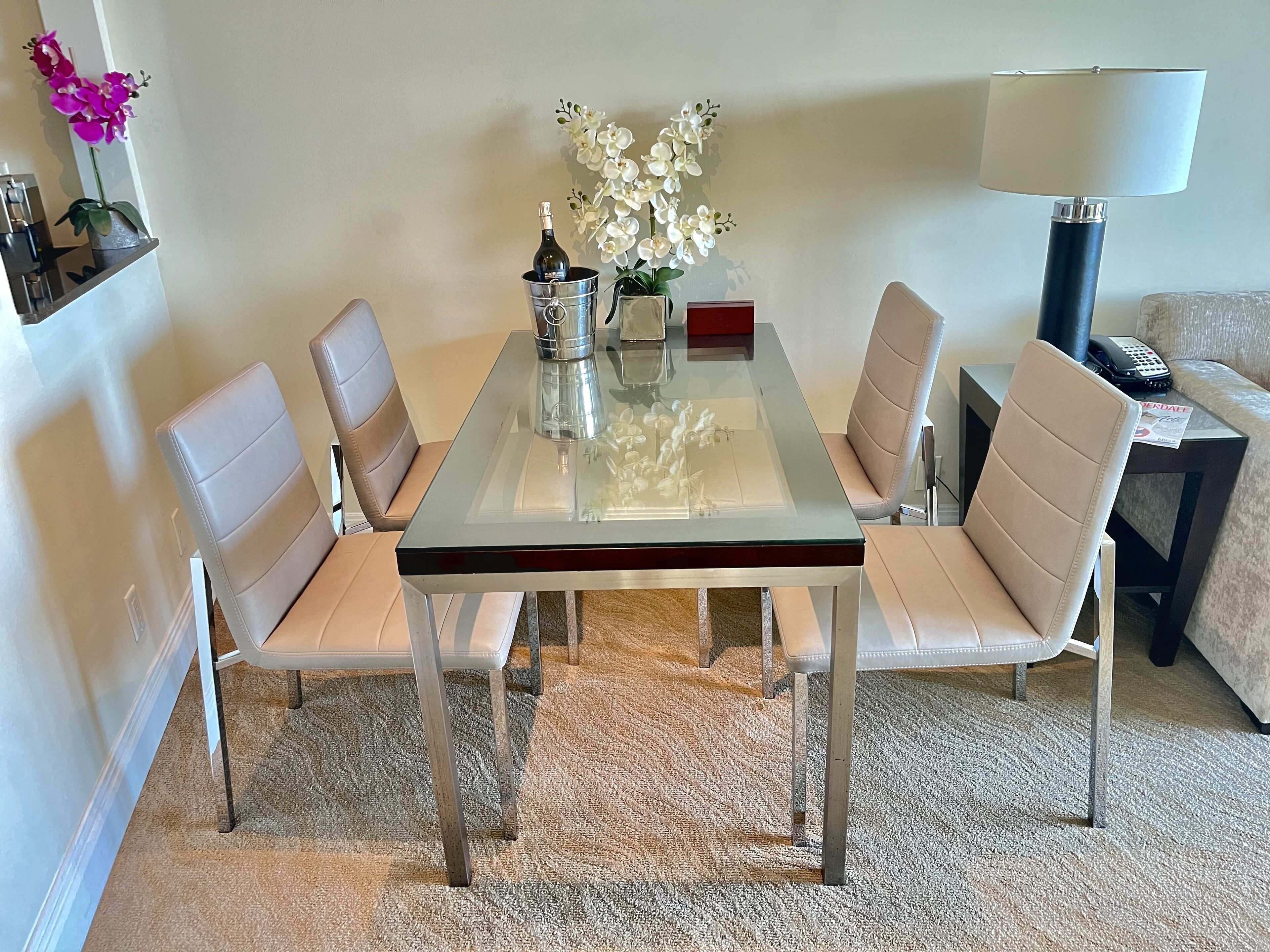 A dining area with a glass-top table, six beige chairs, a vase with white flowers, a bottle of champagne, and a lamp.