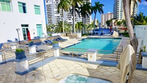 Un espace piscine doté de chaises longues, d’un jacuzzi et offrant une vue sur de hauts immeubles et des palmiers.