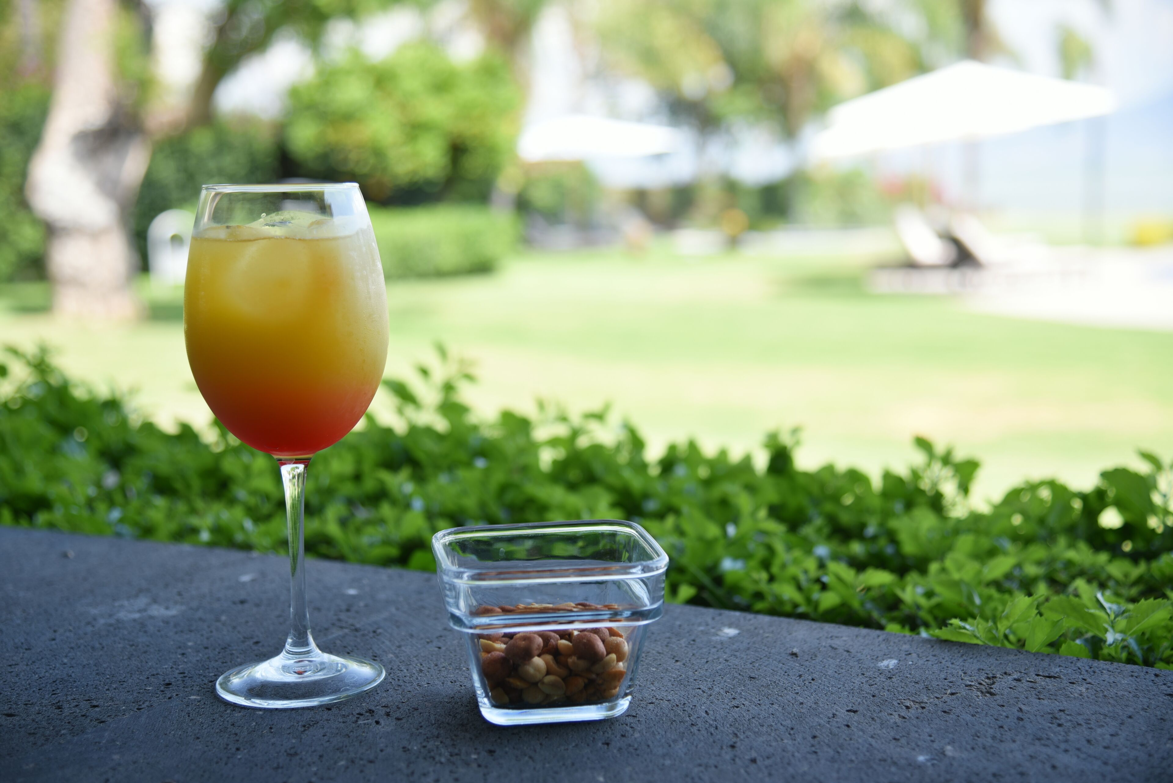 A glass of orange drink on a table with a small container of nuts beside it, set against a blurred outdoor background.