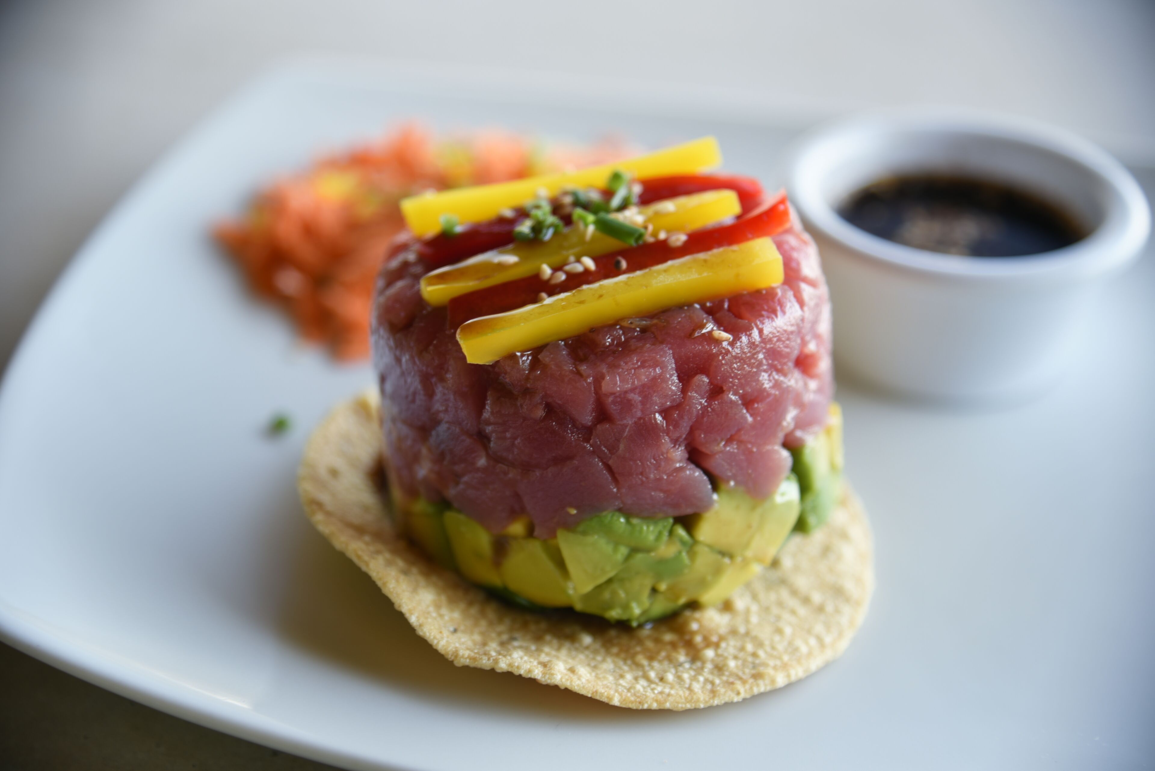Una porzione di tonno fresco a cubetti su un letto di avocado, guarnita con peperoni gialli e rossi, servita con contorno di verdure sottaceto e una piccola ciotola di salsa.