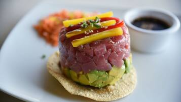 Una porzione di tonno fresco a cubetti su un letto di avocado, guarnita con peperoni gialli e rossi, servita con contorno di verdure sottaceto e una piccola ciotola di salsa.