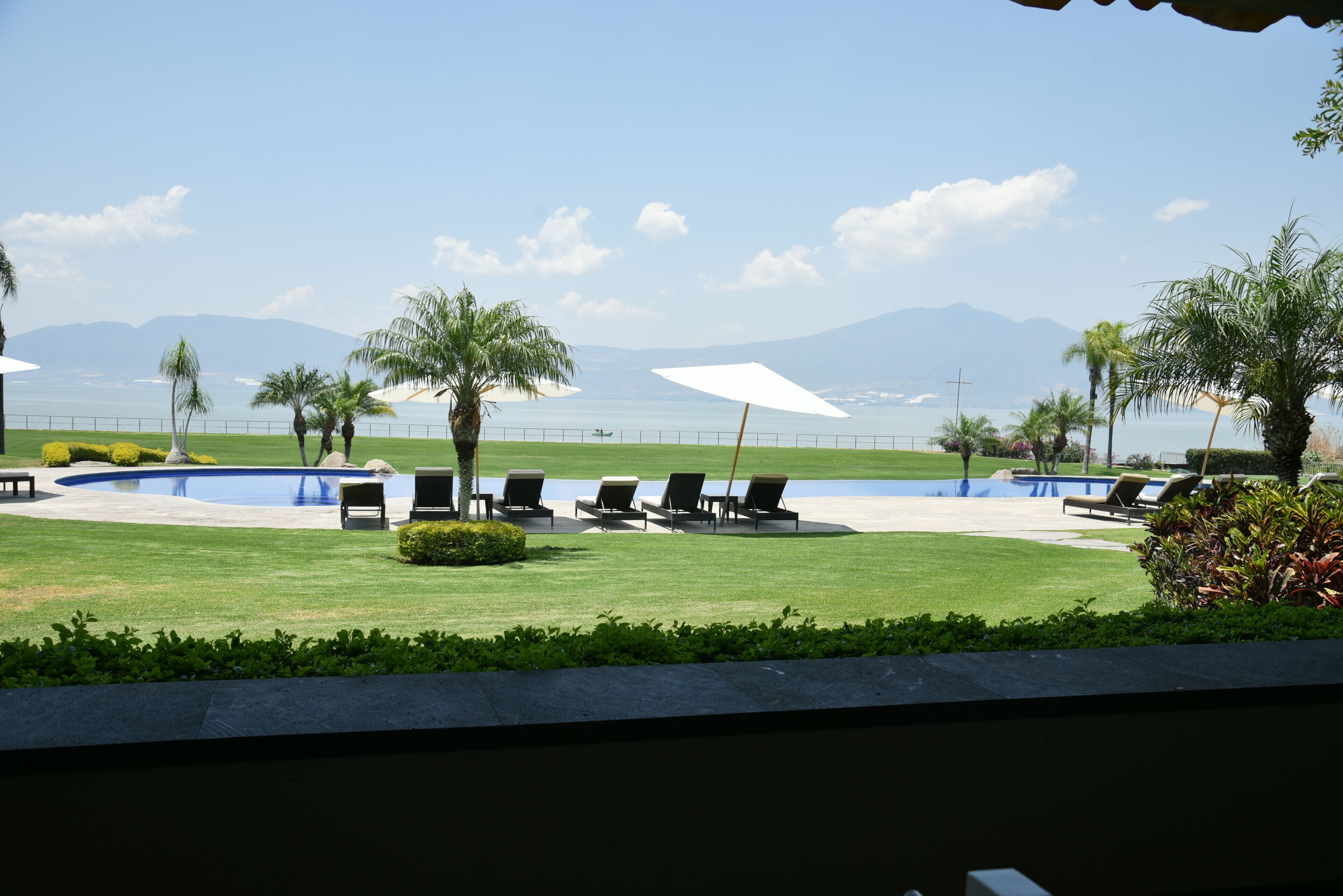 Un área junto a la piscina con sillas de descanso y sombrillas, un césped bien cuidado, y vistas al mar y a las montañas al fondo.