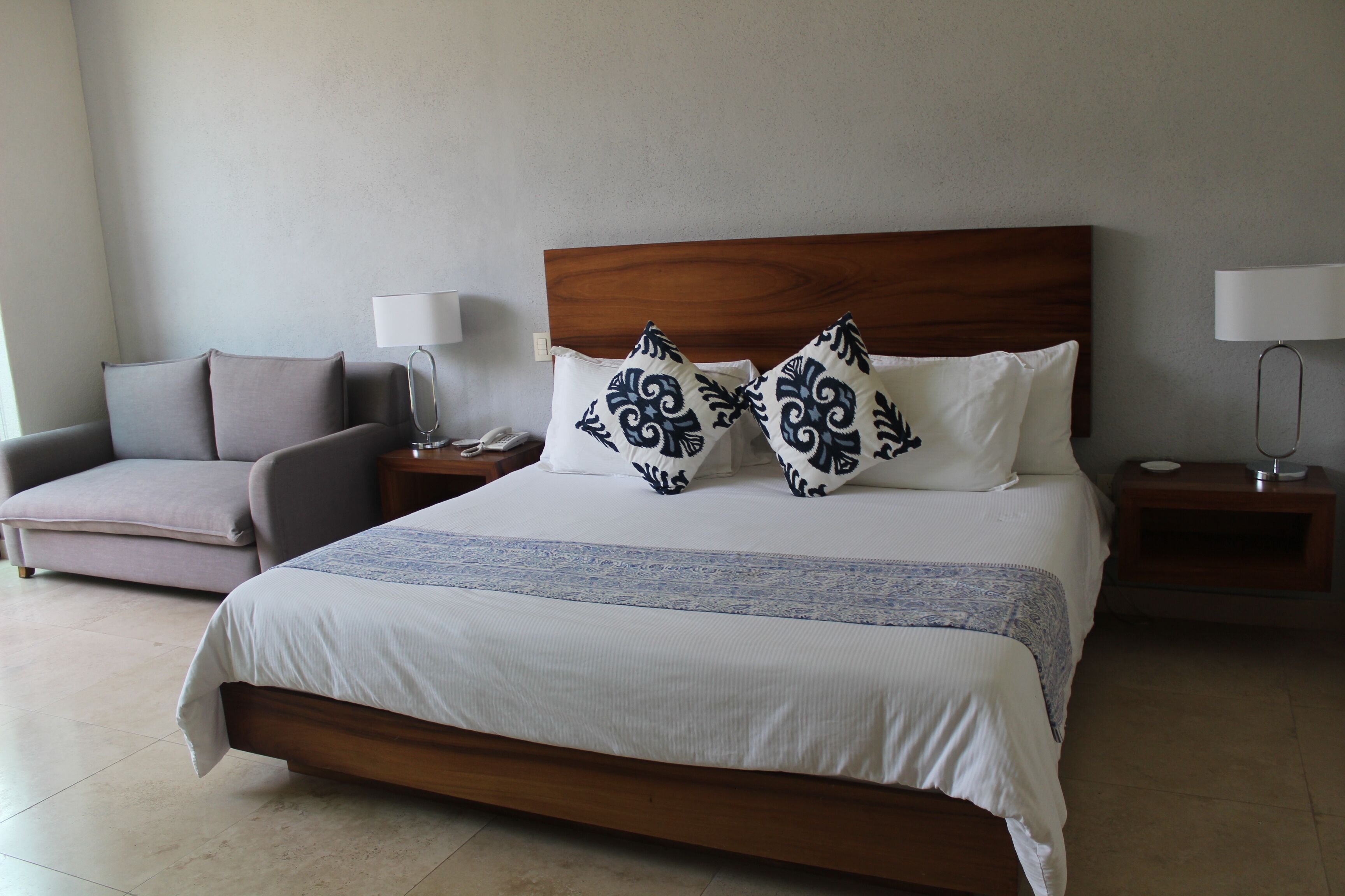 Una habitación de hotel ordenada con una cama grande, un sofá, dos mesitas de noche con lámparas y una lámpara de pared.