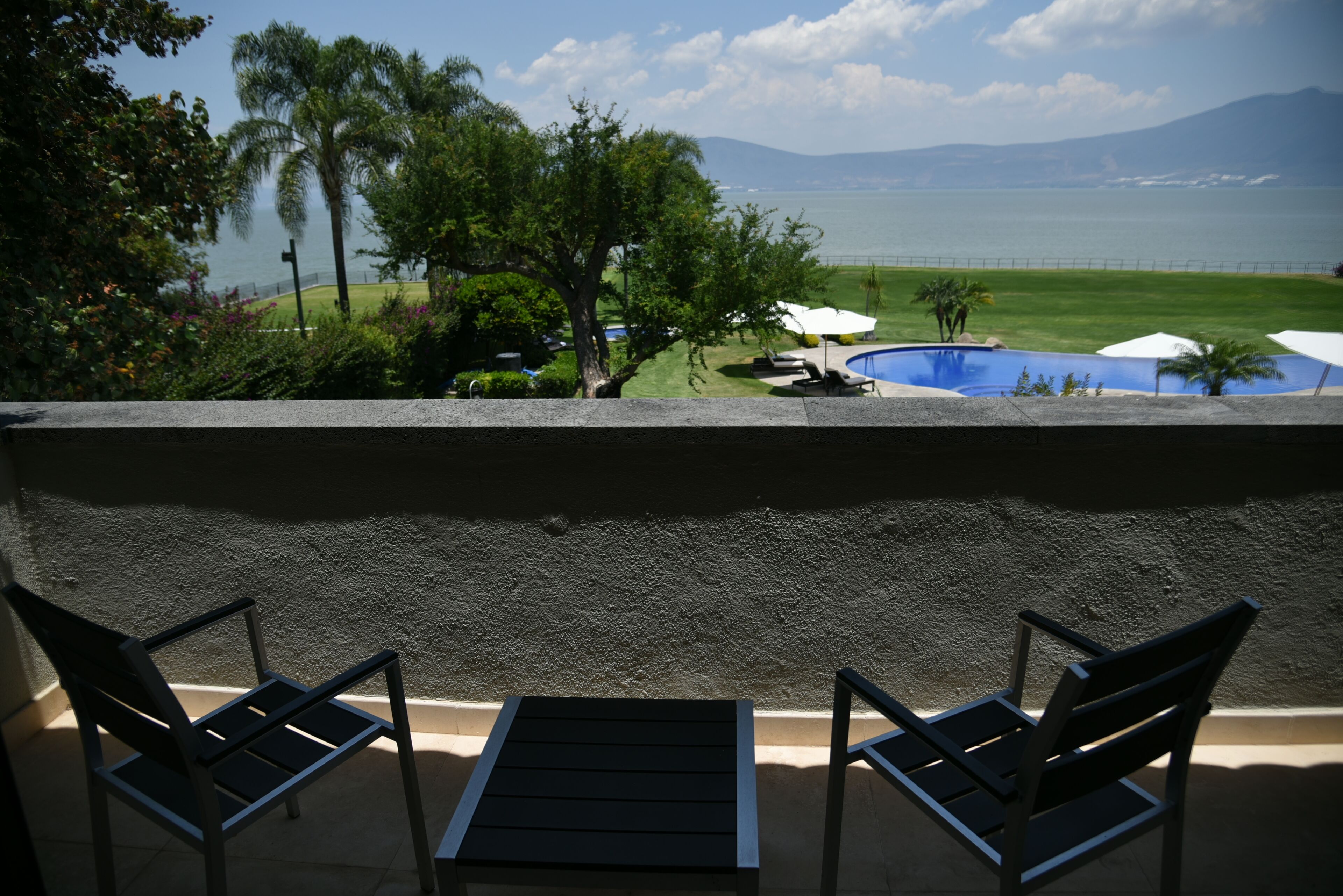 Un balcón con dos sillas y una mesa, con vista a una piscina y al mar y las montañas.
