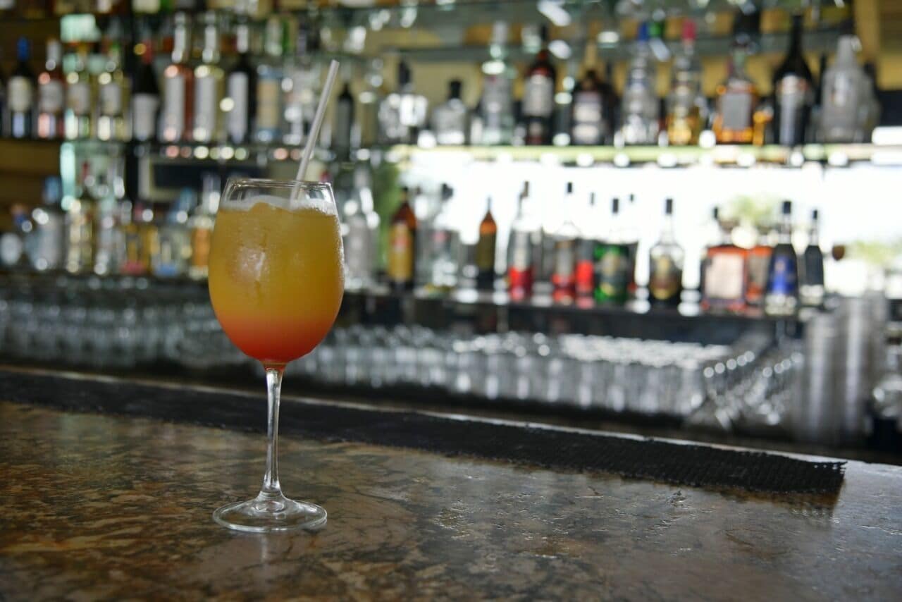 Een cocktailglas met een rietje op een barkruk, met een onscherpe achtergrond van verschillende flessen.