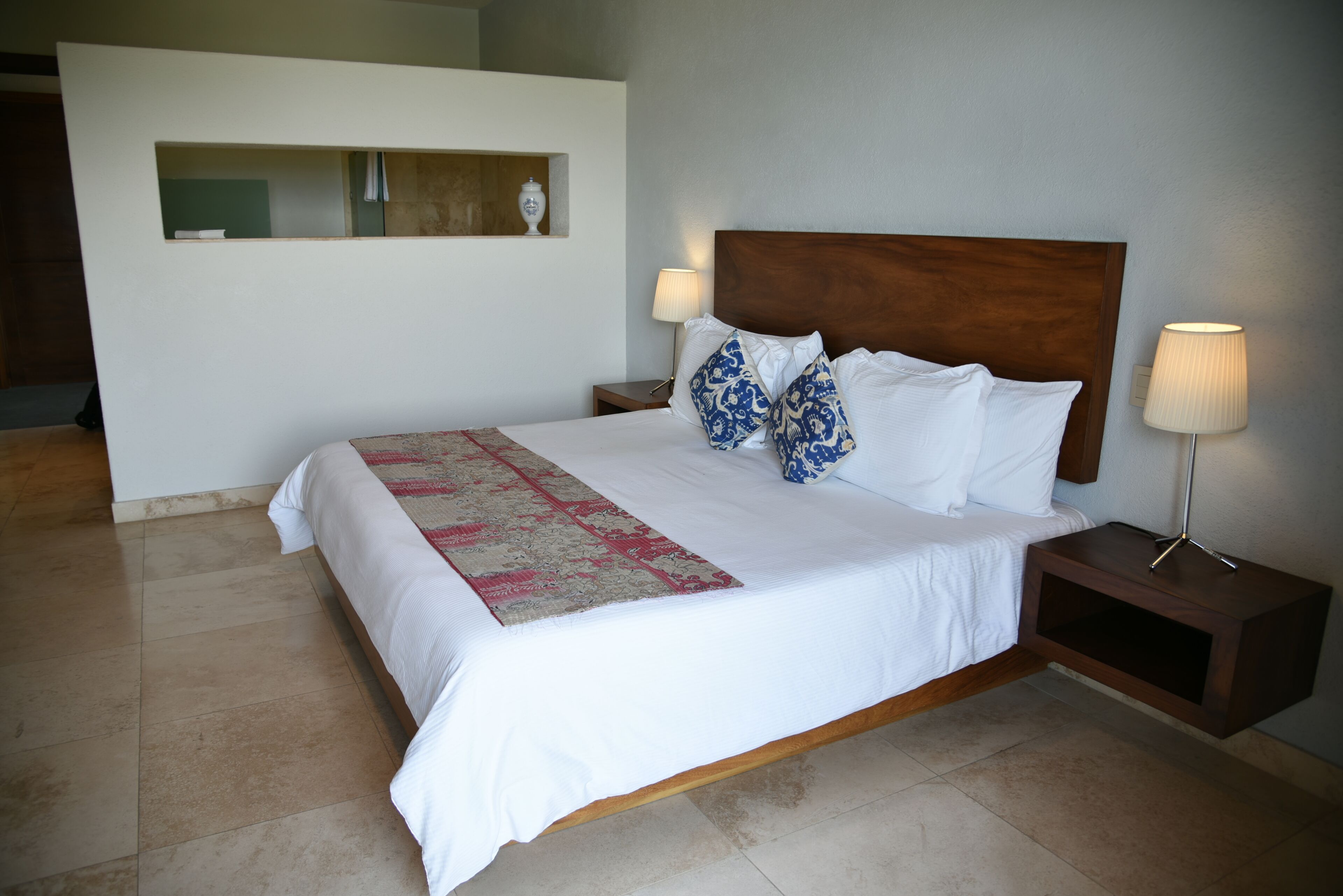 A neatly made bed with white bedding and blue patterned pillows, a bedside table with a lamp, and a small shelf with a vase.