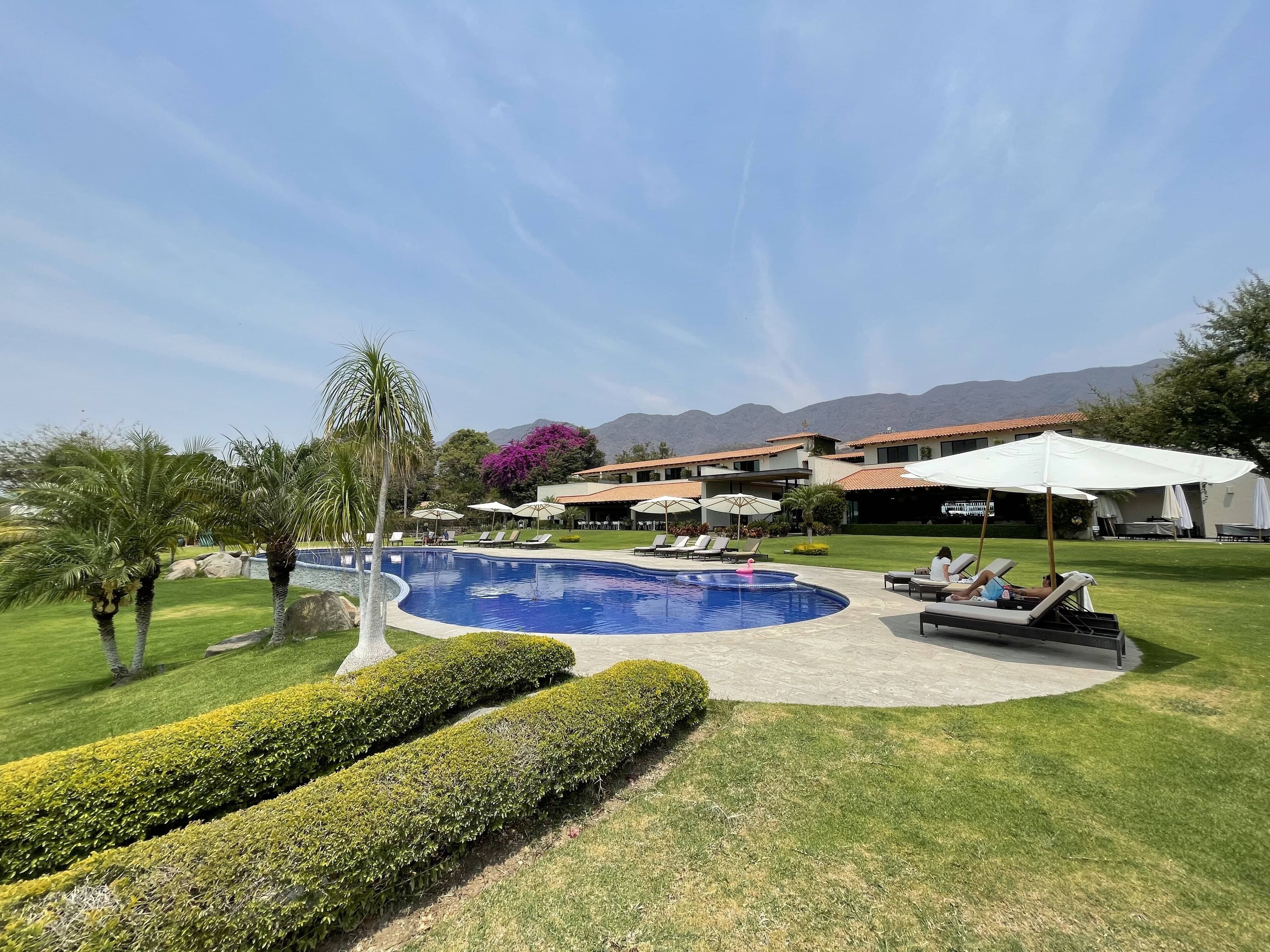 Zona de piscina con sillones y sombrillas, rodeada de césped cuidado y con montañas al fondo.