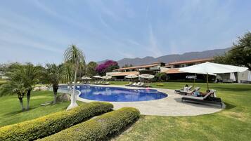 Zona de piscina con sillones y sombrillas, rodeada de césped cuidado y con montañas al fondo.