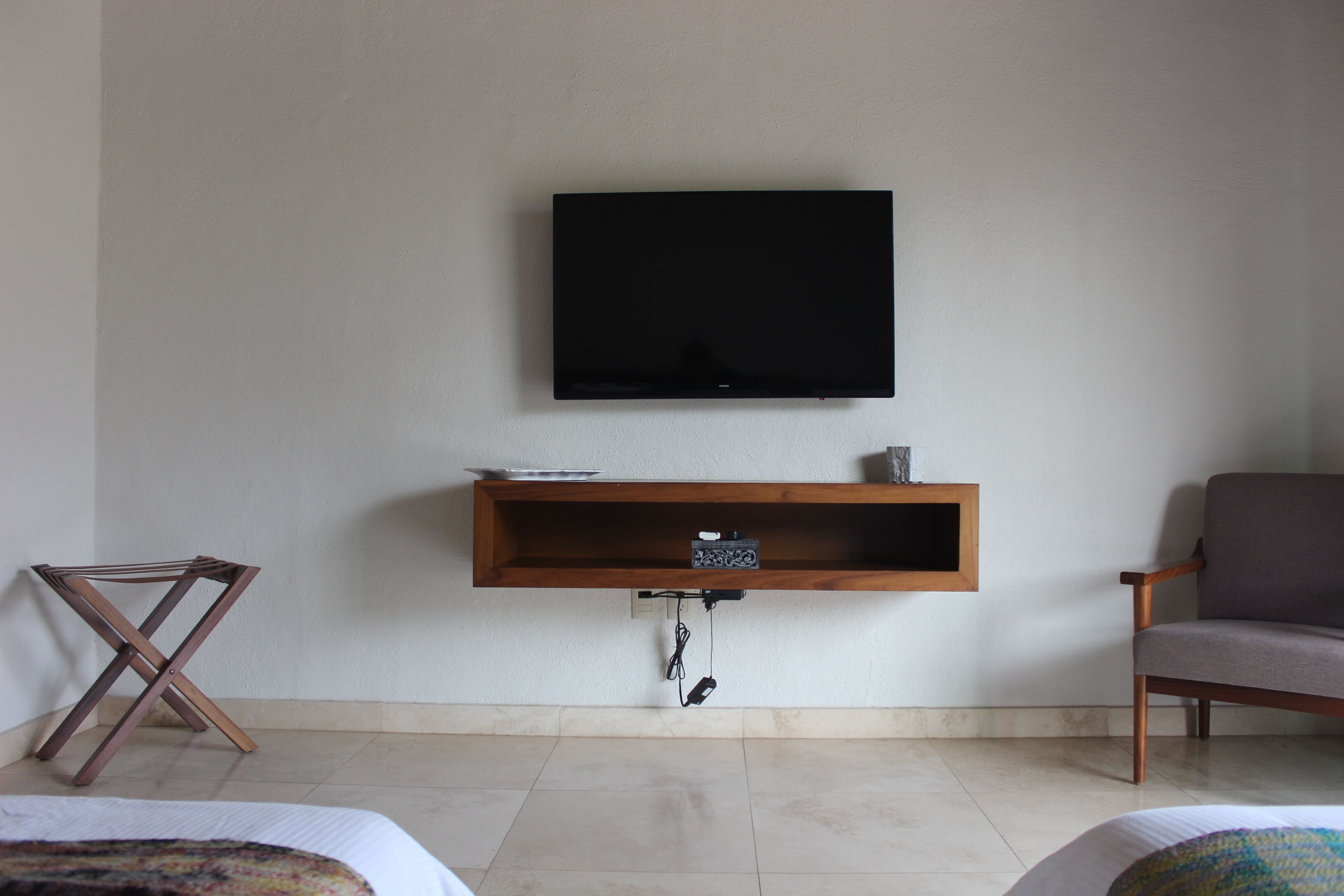 Una habitación con una televisión de pantalla plana montada en la pared, un estante de madera debajo, una silla plegable y una cama con una manta colorida.