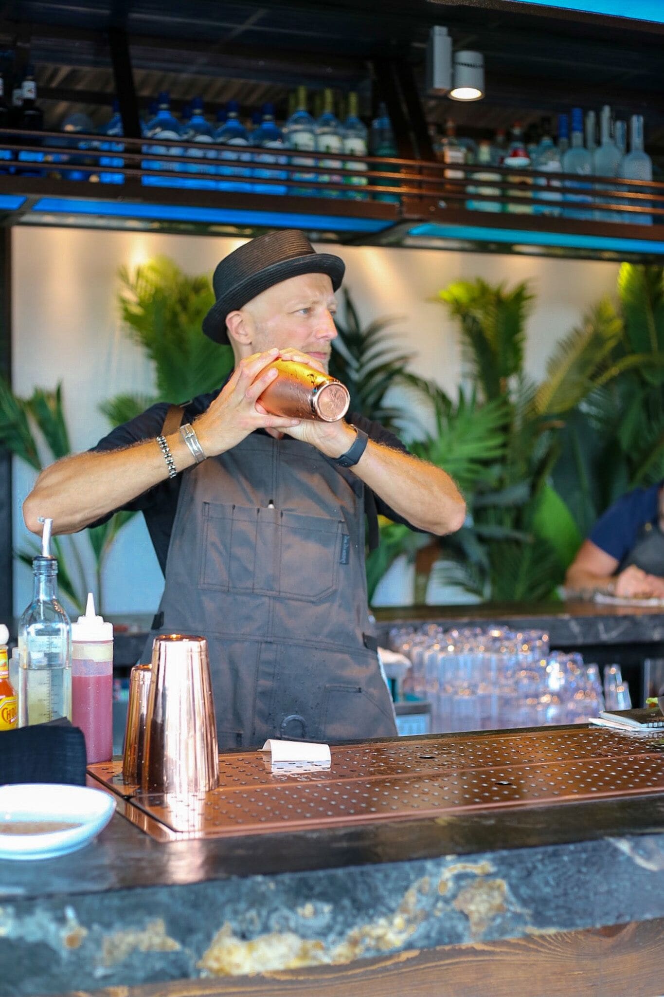 A bartender preparing a drink behind a bar with various bottles and a copper shaker.