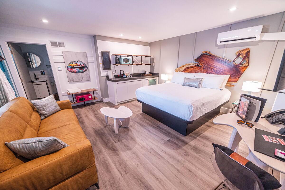 Ein modernes Hotelzimmer mit einem großen Bett, einer Couch, einem kleinen Tisch, einem Schreibtisch mit Stuhl, einer Küchenzeile und einem Badezimmer.