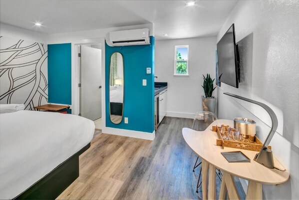 Une chambre d’hôtel moderne équipée d’un lit, d’un petit bureau avec une lampe, d’un téléviseur fixé au mur et d’une fenêtre donnant sur un paysage verdoyant.