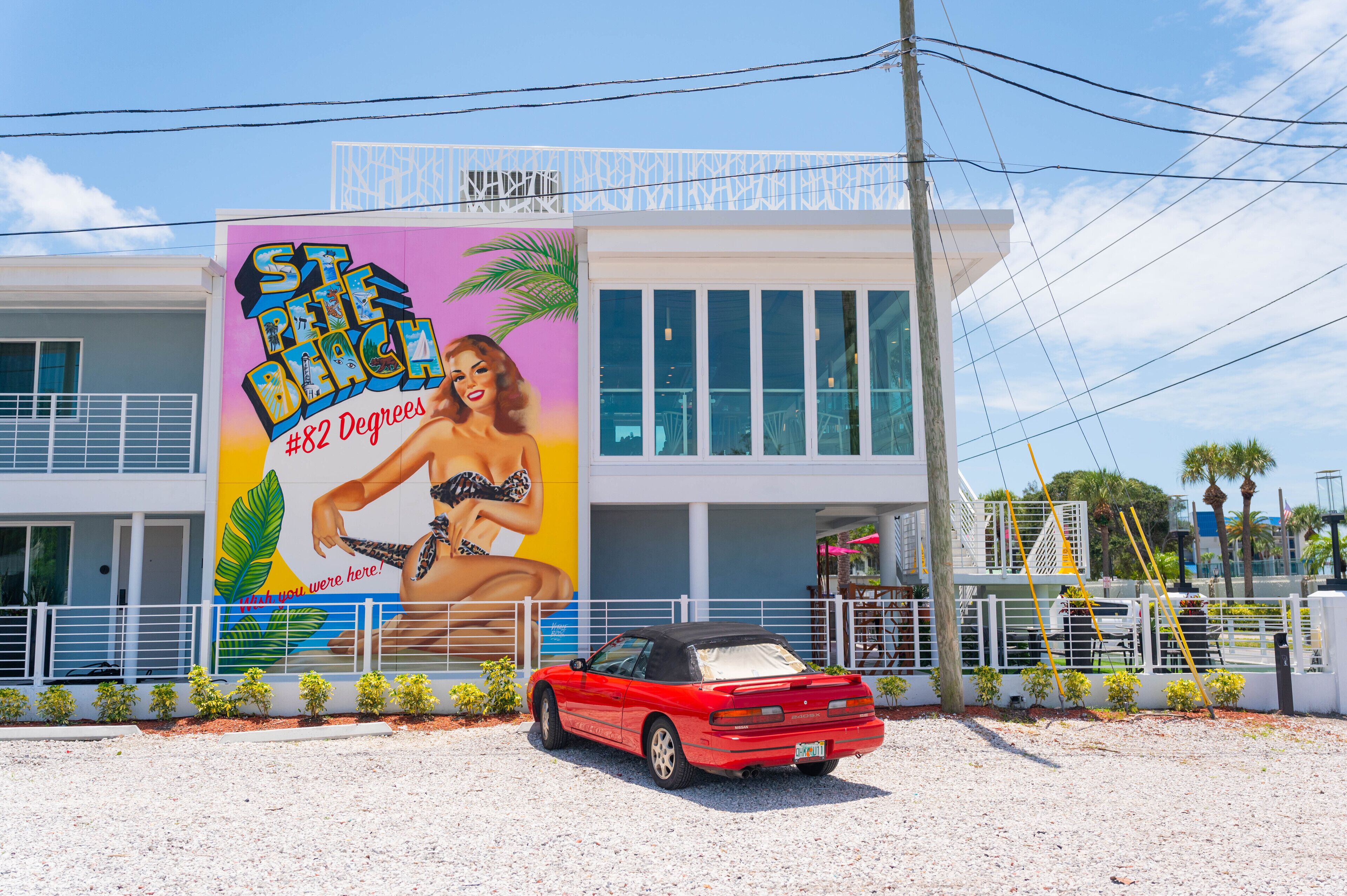 A red convertible parked in front of a building with a large mural advertising a beach destination.