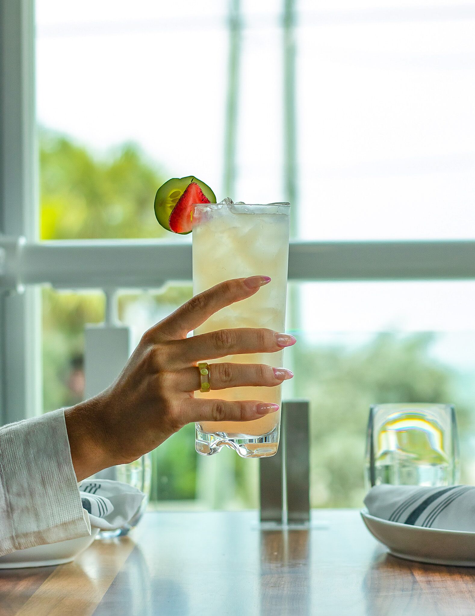 A hand holding a tall glass with a lime wedge and a strawberry slice, set against a window with a blurred outdoor view.