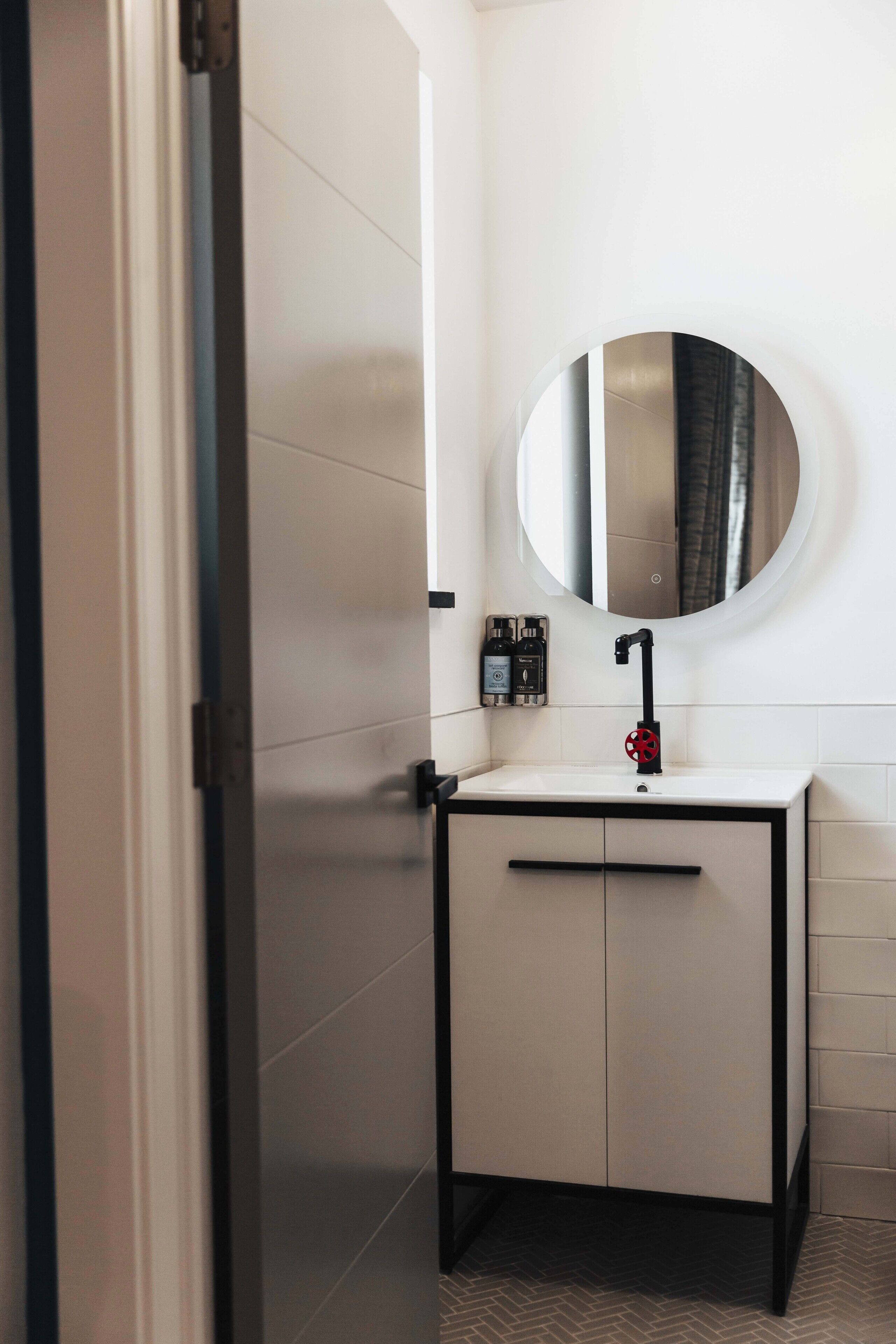 A bathroom with a white sink, black faucet, and a round mirror. There are two bottles of soap on the wall.