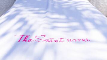 Una cama blanca con un almohadón rosa y el texto "The Saint Hotel" sobre una superficie blanca.