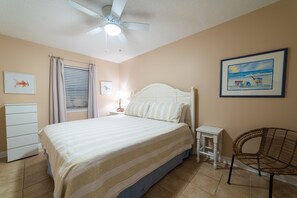 A bedroom with a bed, a ceiling fan, a window with curtains, a nightstand, a chair, and a framed picture on the wall.