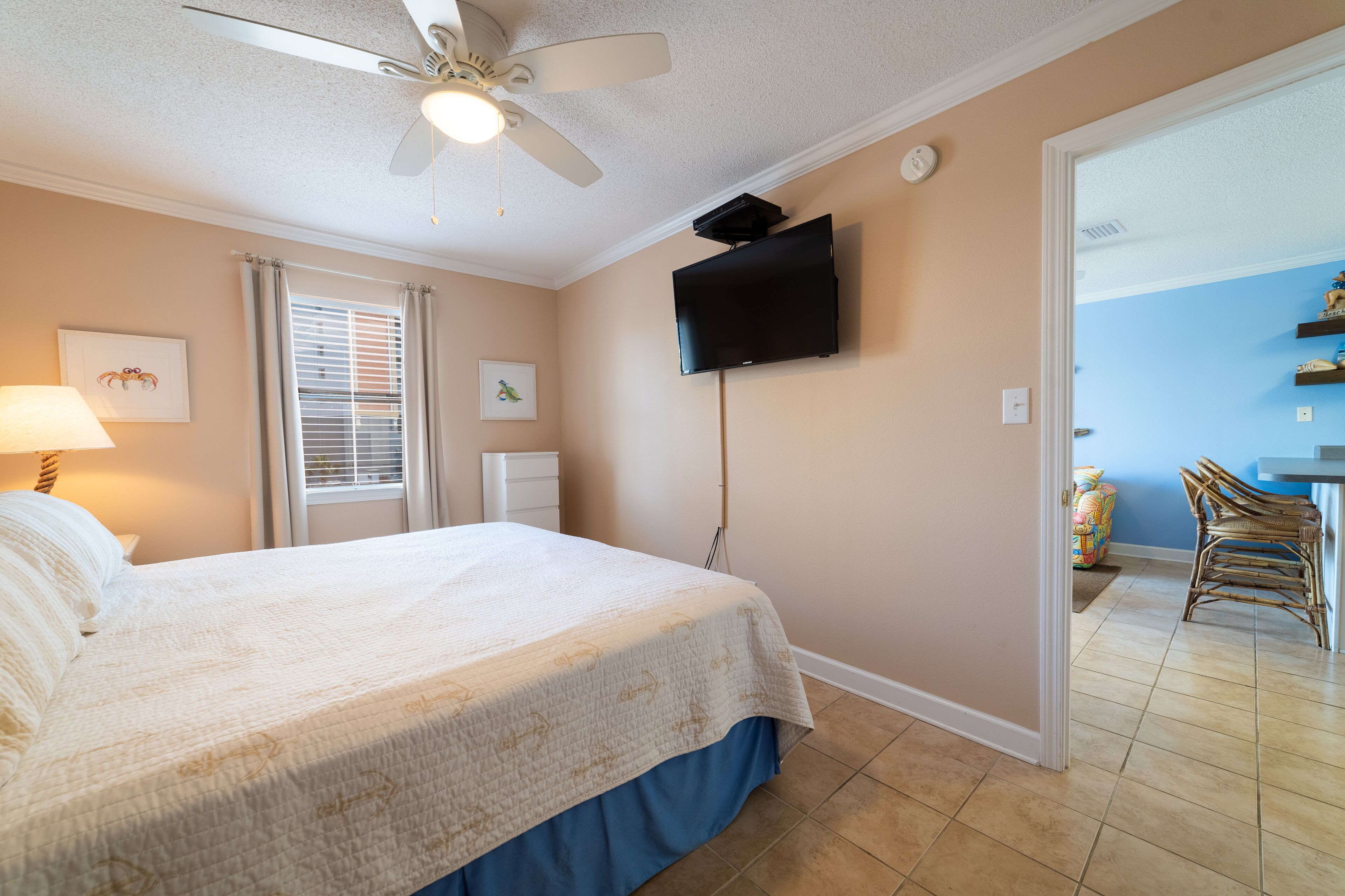 A bedroom with a bed, ceiling fan, TV, and a view into the dining area.