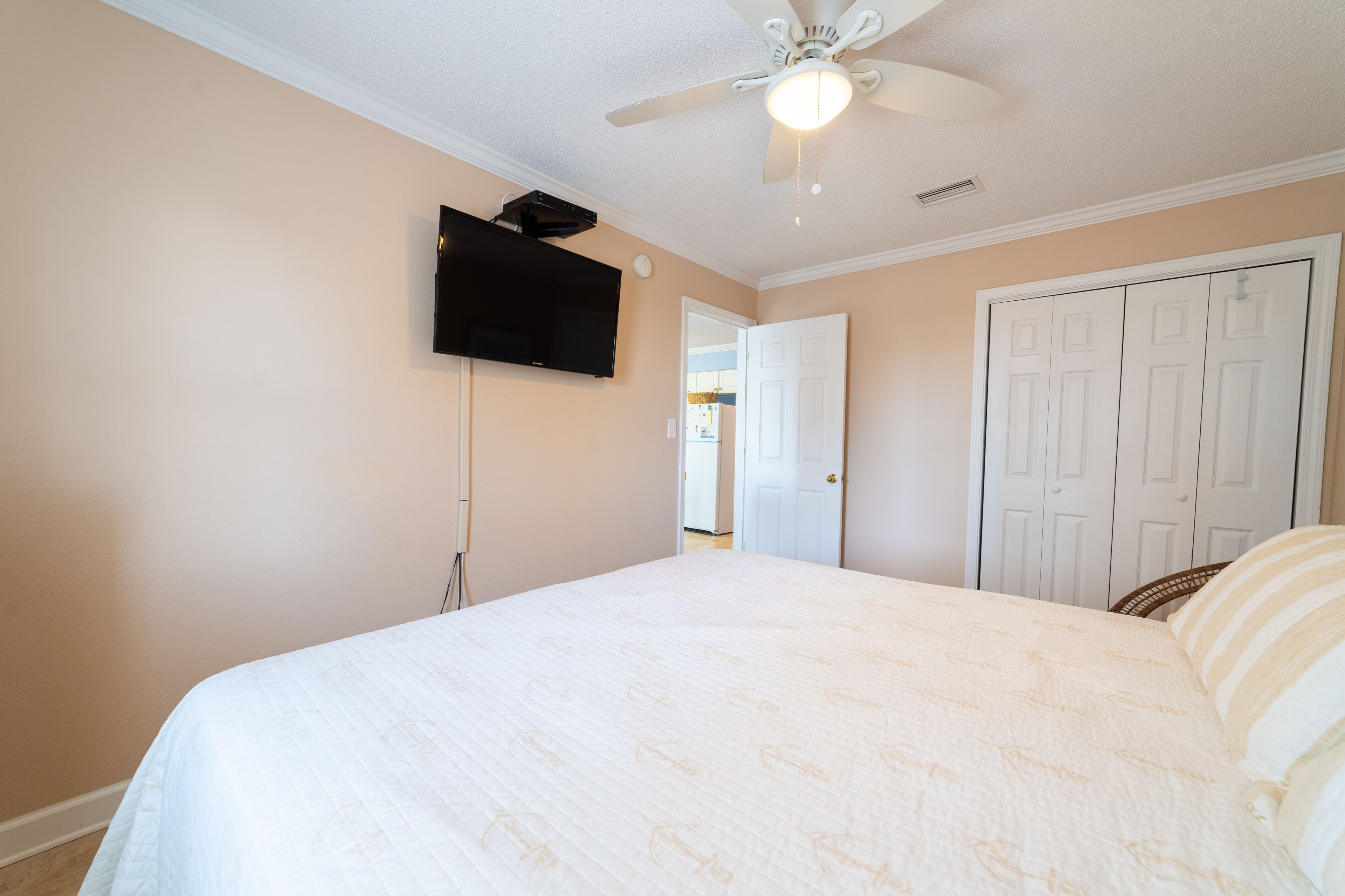 A bedroom with a bed, a ceiling fan, a television mounted on the wall, and a white door.