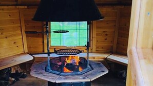 Puinen mökki sisältä, keskellä on puukamiina, ikkunasta avautuu näkymä vehreään maisemaan ja vasemmalla puolella on penkki.