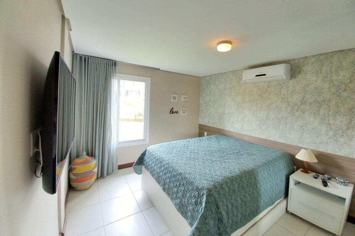 A hotel room with a bed, a TV, a window with curtains, a wall-mounted air conditioner, and a bedside table with a lamp.