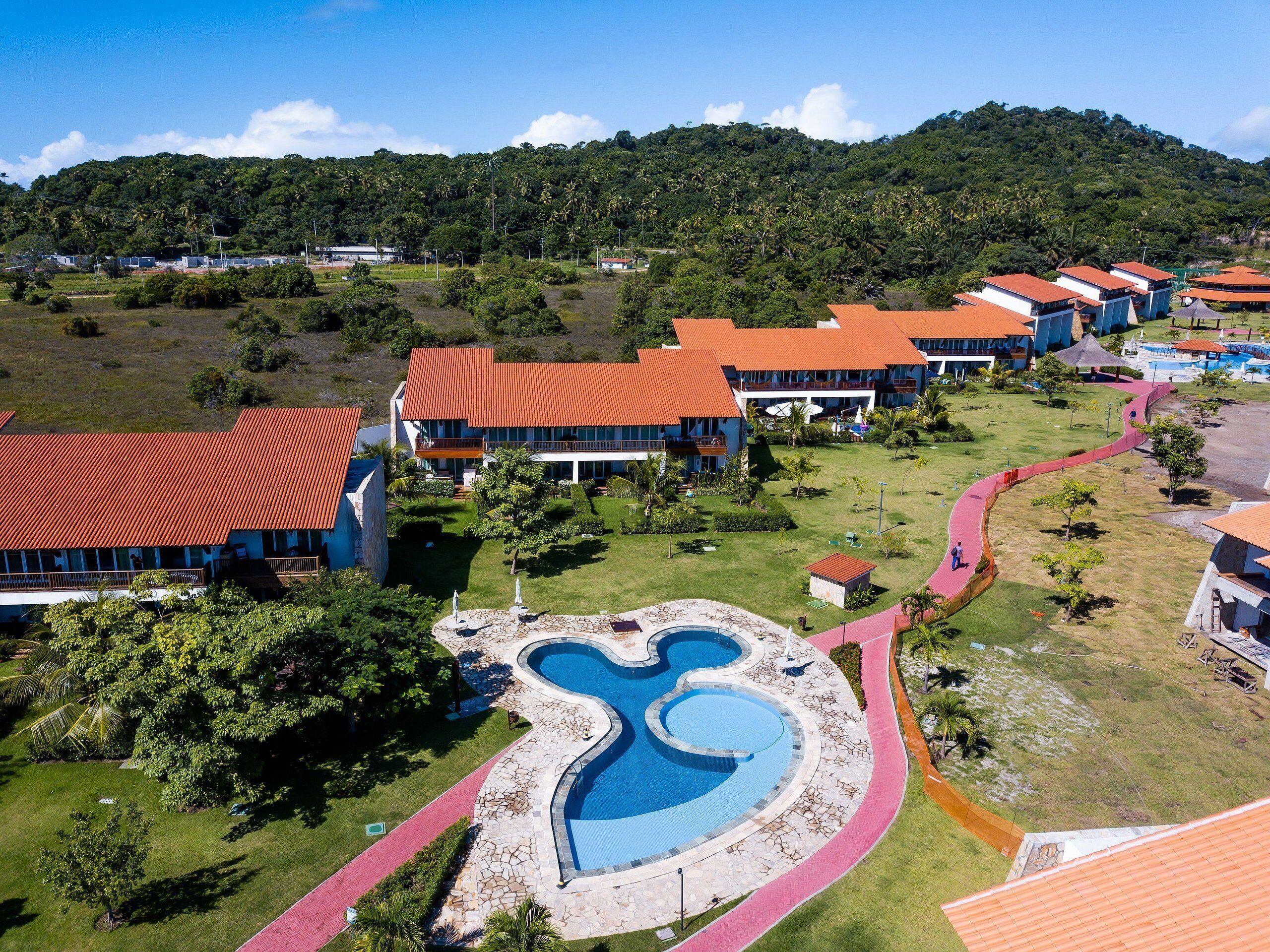 A resort with multiple buildings, a central swimming pool, and a mountainous backdrop.