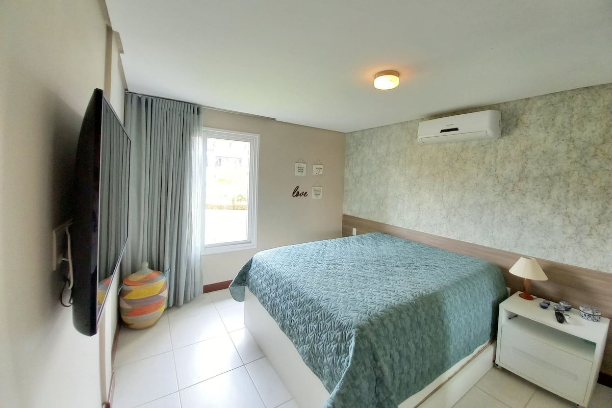 A hotel room with a bed, a TV, a window with curtains, a wall-mounted air conditioner, and a bedside table with a lamp.
