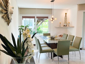 A dining area with a long table, chairs, a chandelier, and a view of greenery outside.