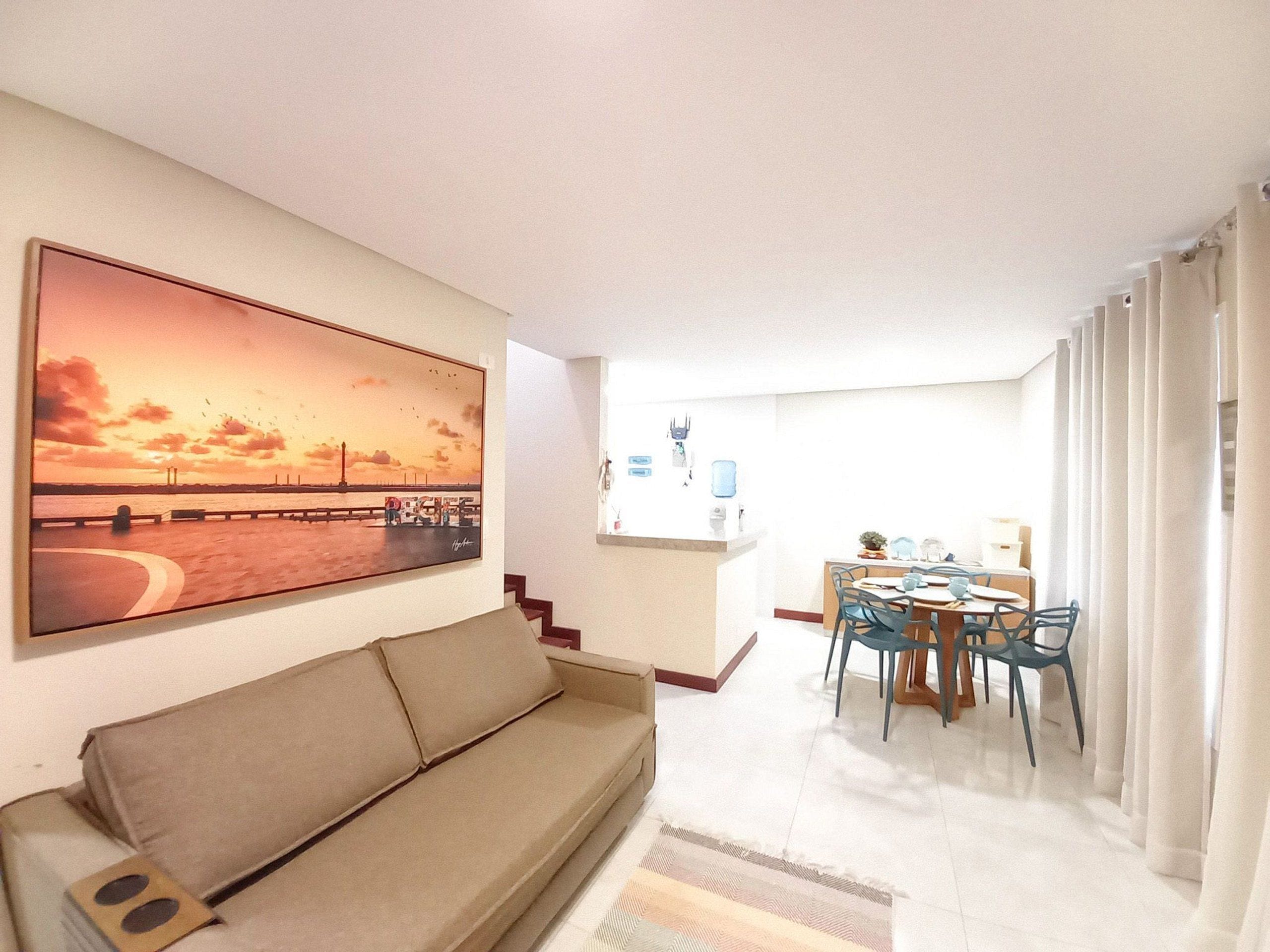 Uma sala de estar com sofá, um espaço de refeições com cadeiras e mesa, um quadro na parede e vista para o exterior.