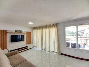 Ein Wohnzimmer mit Flachbildfernseher, einer Couch, einem Fenster mit Vorhängen und einem sichtbaren Balkon durch das Fenster.