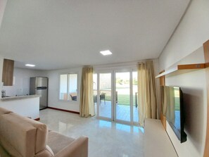 Un salón moderno con televisor de pantalla plana, un sofá, una zona de cocina y un ventanal corredero que da a un patio.