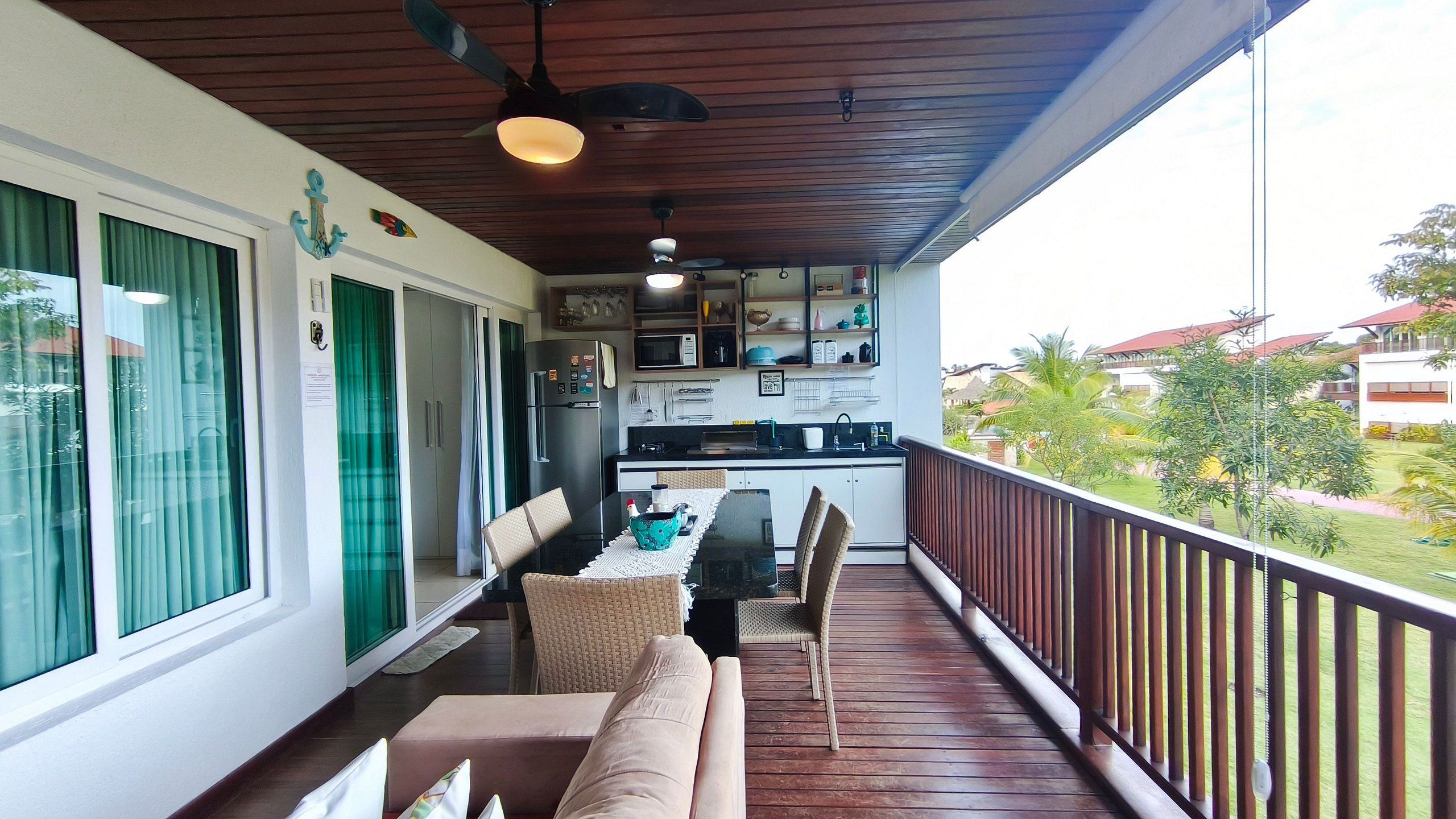 A balcony with a wooden floor, a dining table with chairs, a kitchen area with appliances, and a view of the surrounding landscape.